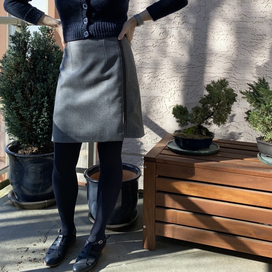 Standing with hands on hips wearing grey mini skirt, blue rights and blue shoes, shown from the waist down. Two small bonsai trees and a dwarf cypress behind.